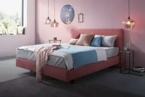 Stilvolles Schlafzimmer mit einem roten Polsterbett, dekoriert mit bunten Kissen und modernen Nachttischen, rosa Wände und elegante Hängeleuchten.